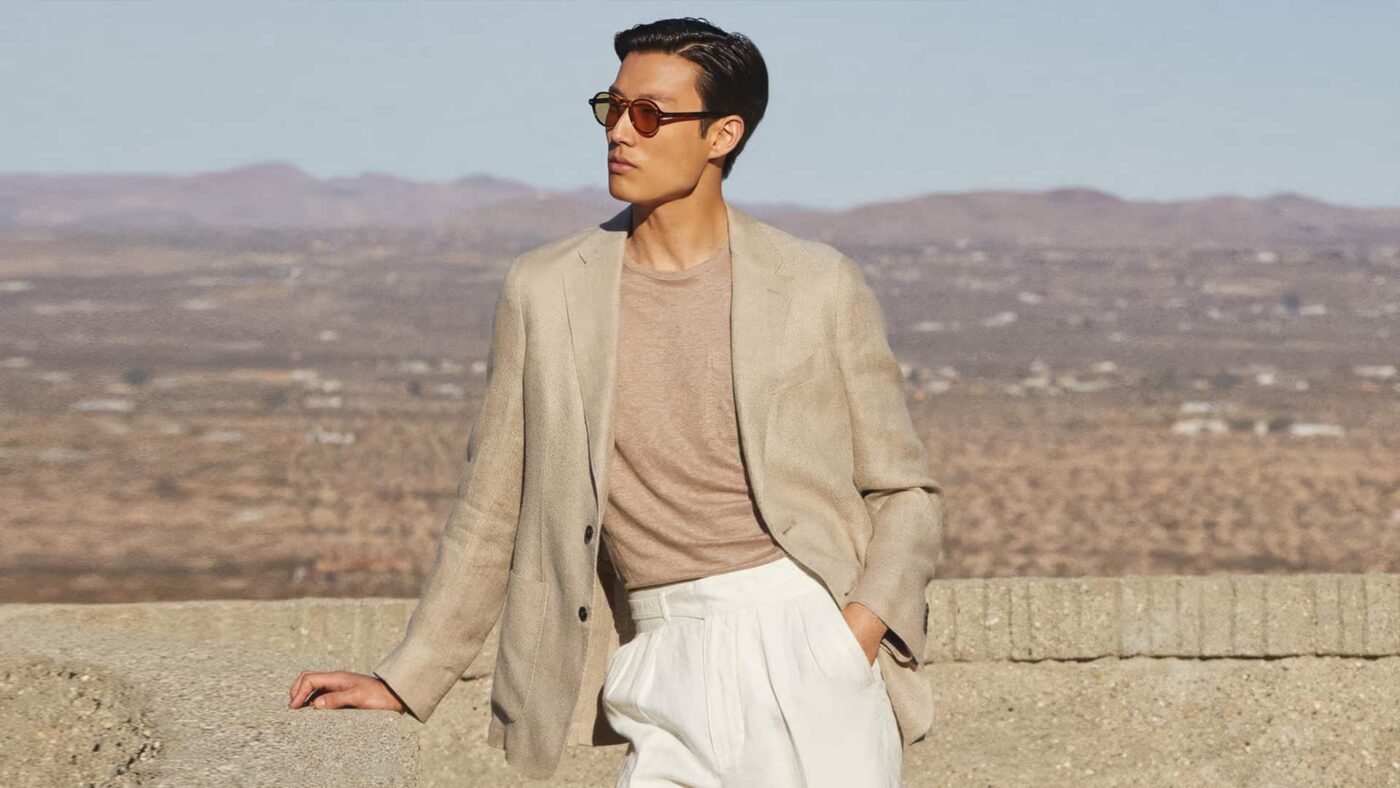 A male model wearing a beige linen blazer, light brown tee, and white trousers with shades. 