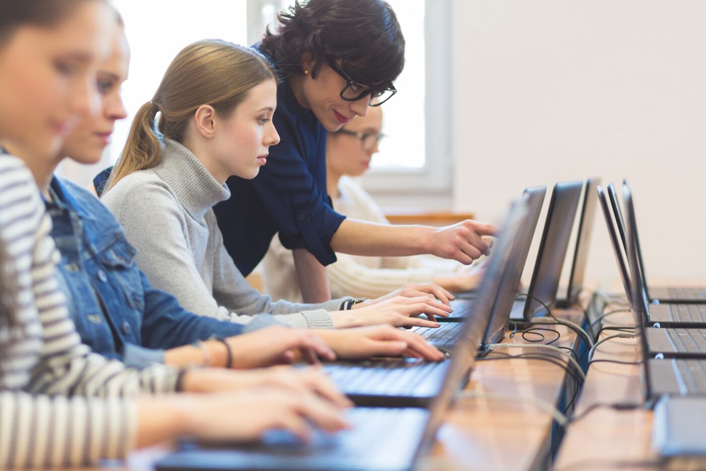 Side view of group of female students coding on laptops in a computer lab. Teacher pointing at the computer screen.