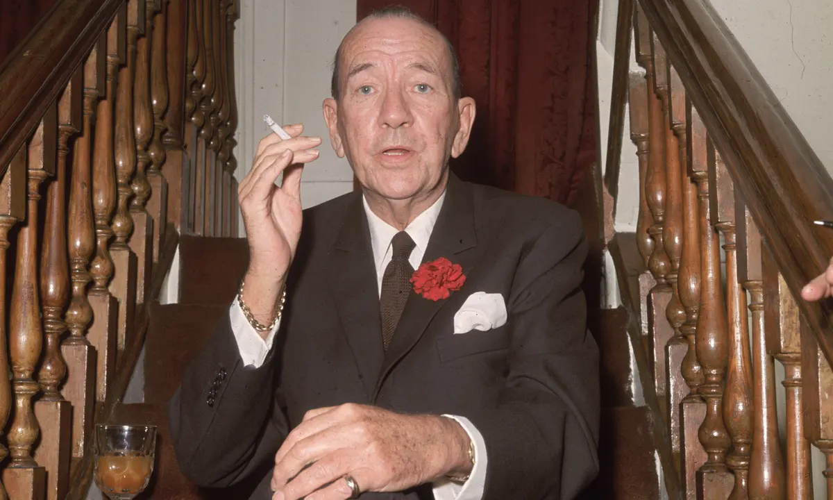 Noel Coward holding a cigarette, sitting on a wooden staircase. 